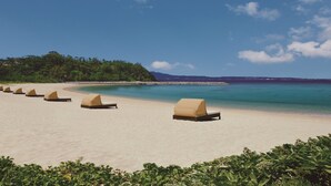A beach with six sun loungers aligned along the shore, a clear blue sky, and a forested hill in the background. - The Terrace Club Wellness Thalasso At Busena (Nago)
