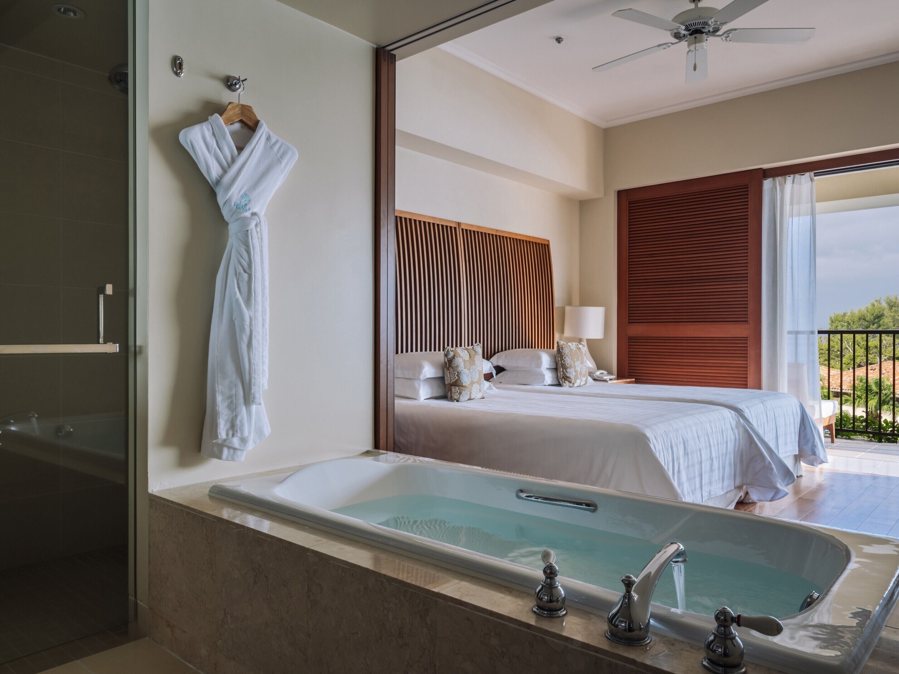 A modern hotel room with a large bathtub, a neatly made bed, and a balcony with a view.