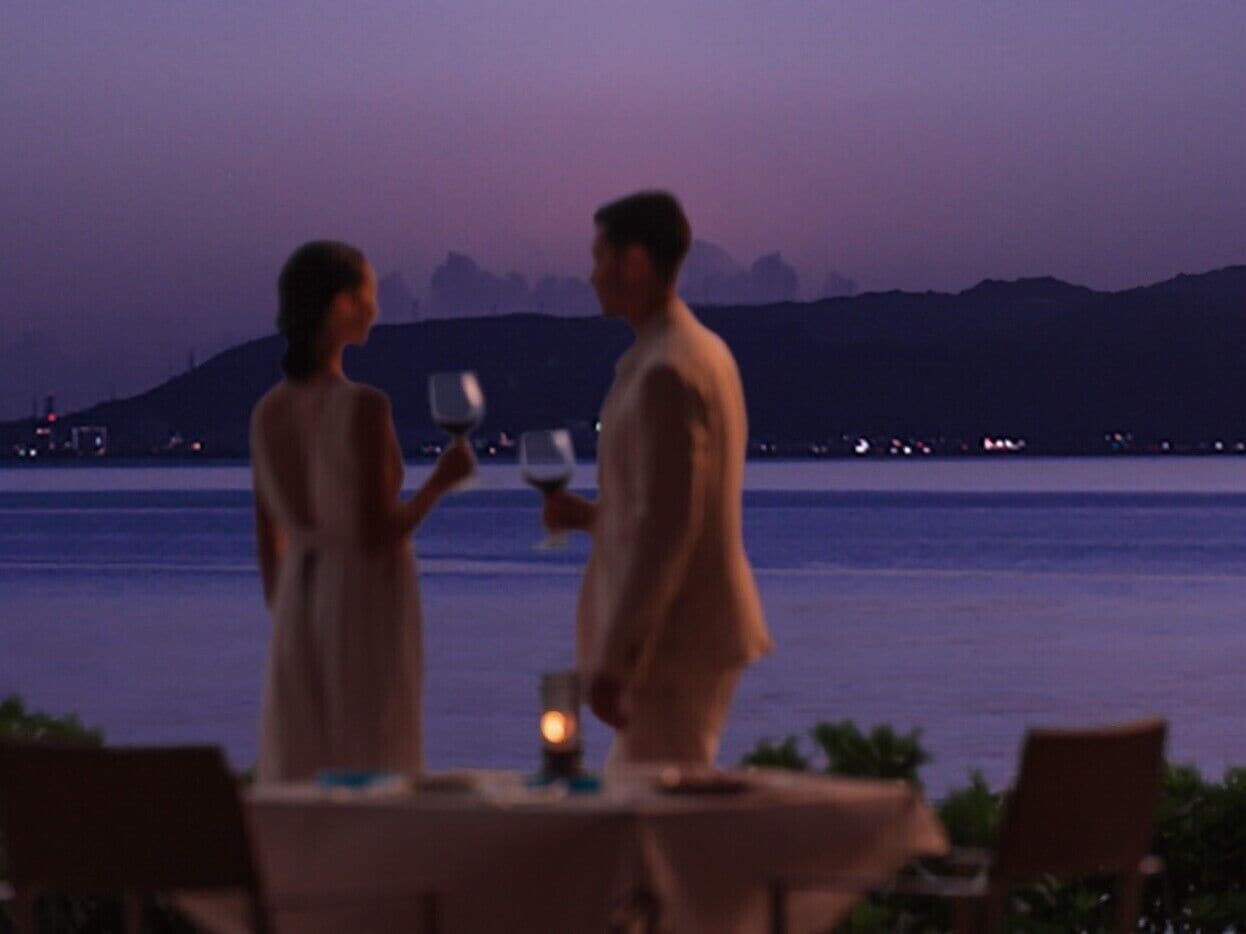A couple dining at a table with a view of the sea and mountains in the background.