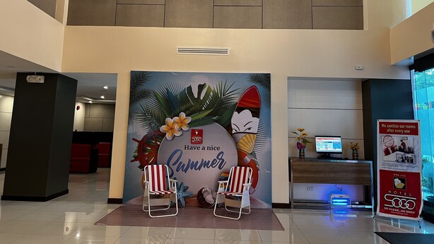 Zona de recepción de hotel con un mural de temática tropical, dos sillas de playa, un mostrador de recepción y un cartel sobre la sanitización de habitaciones.