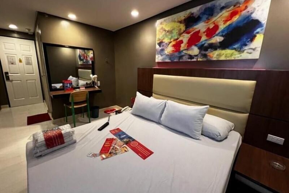 Una habitación de hotel con cama, escritorio, silla, espejo y un cuadro en la pared.