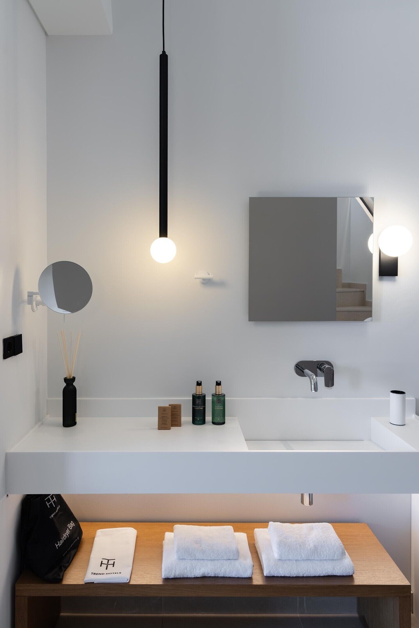 Un baño moderno con tocador blanco, espejo redondo, lámpara colgante negra y banco de madera con toallas.
