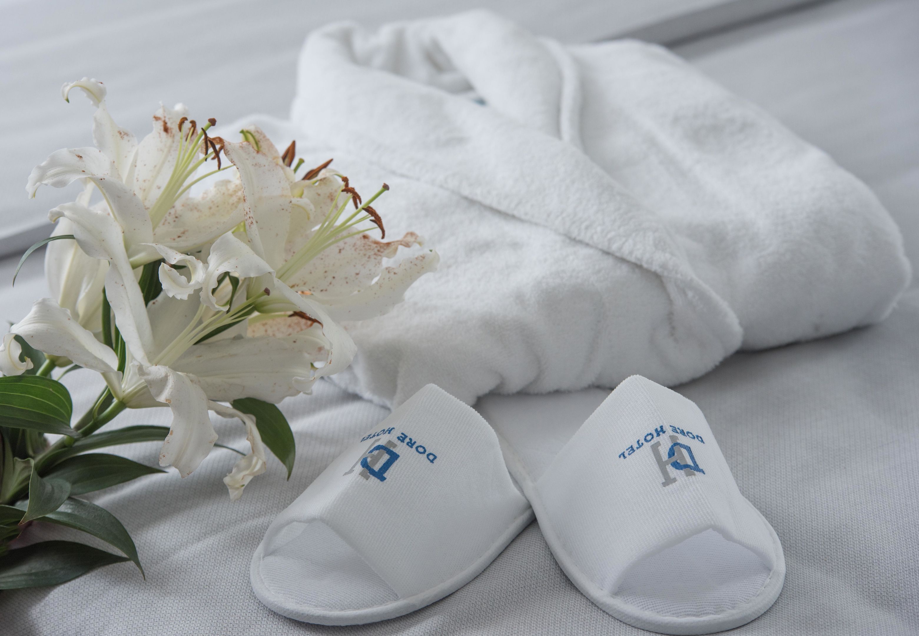 Un par de pantuflas blancas con un logo, una toalla blanca y una flor blanca sobre una cama.