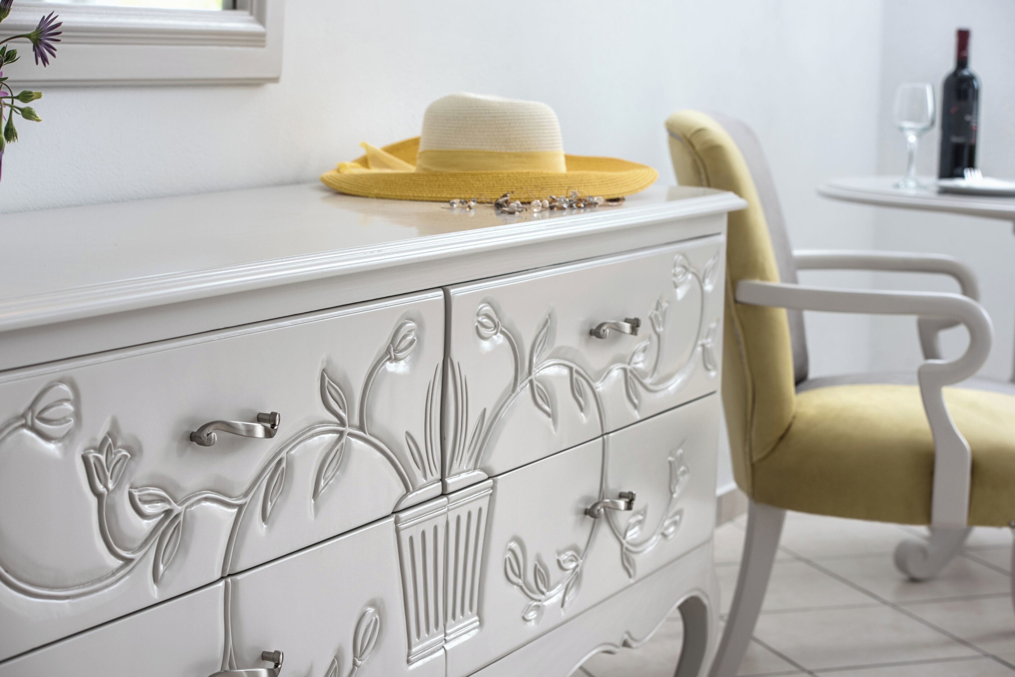 Un aparador blanco con detalles florales, un sombrero amarillo encima y una silla amarilla a su lado.