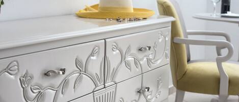 Un aparador blanco con detalles florales, un sombrero amarillo encima y una silla amarilla a su lado.