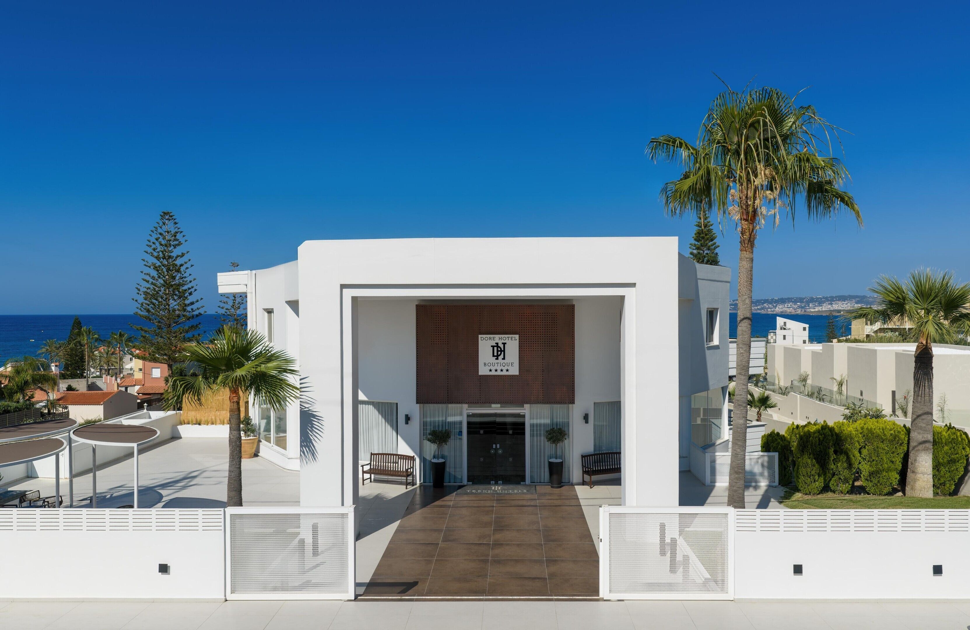 Une entrée d’hôtel moderne offrant une vue dégagée sur la mer, une enseigne indiquant le nom de l’hôtel et un chemin pavé menant à l’entrée.