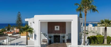 Una entrada de hotel moderna con vista despejada al mar, un letrero con el nombre del hotel y un camino pavimentado que lleva a la entrada.