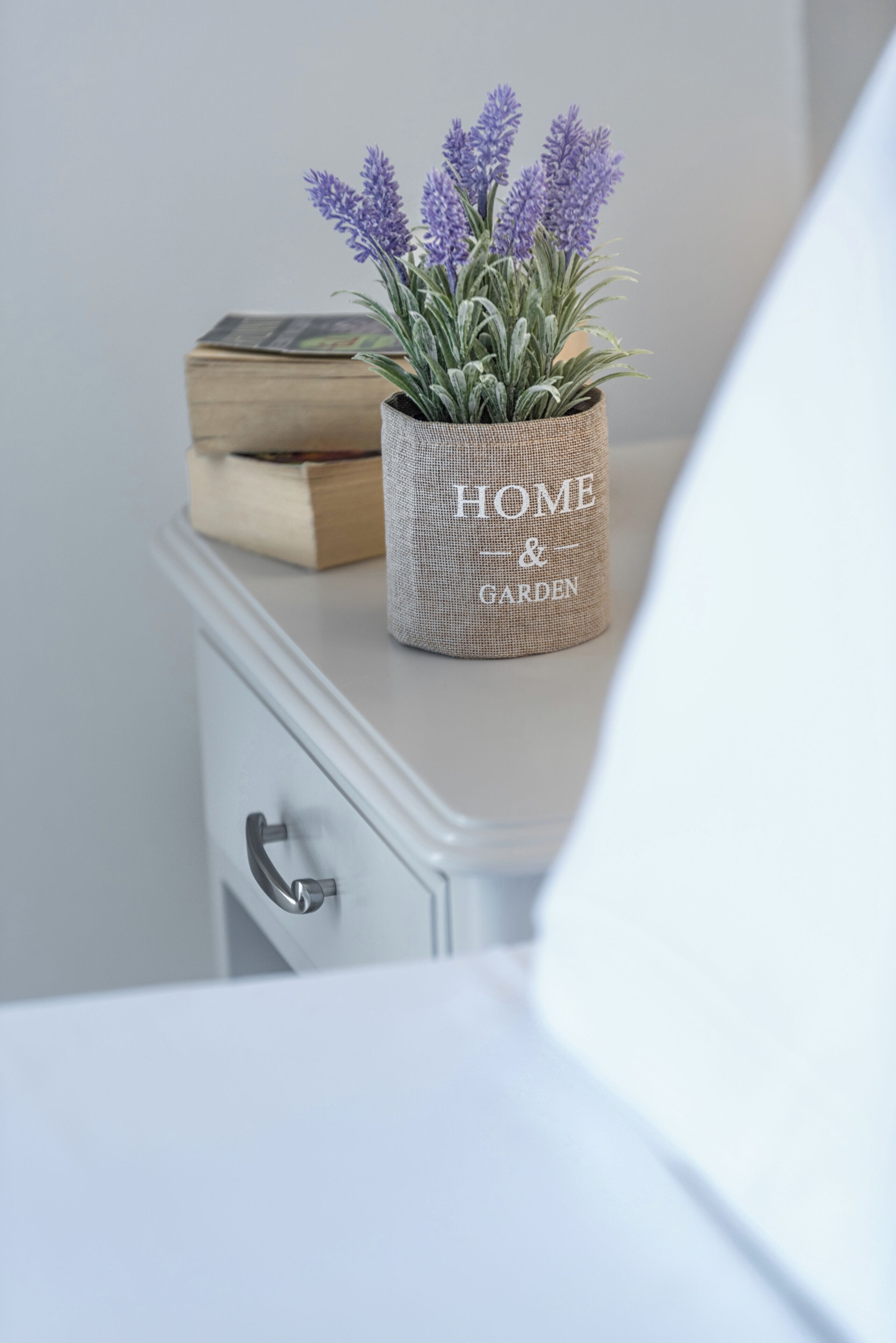 Une table de chevet blanche avec un panier en osier contenant des plants de lavande et un livre.