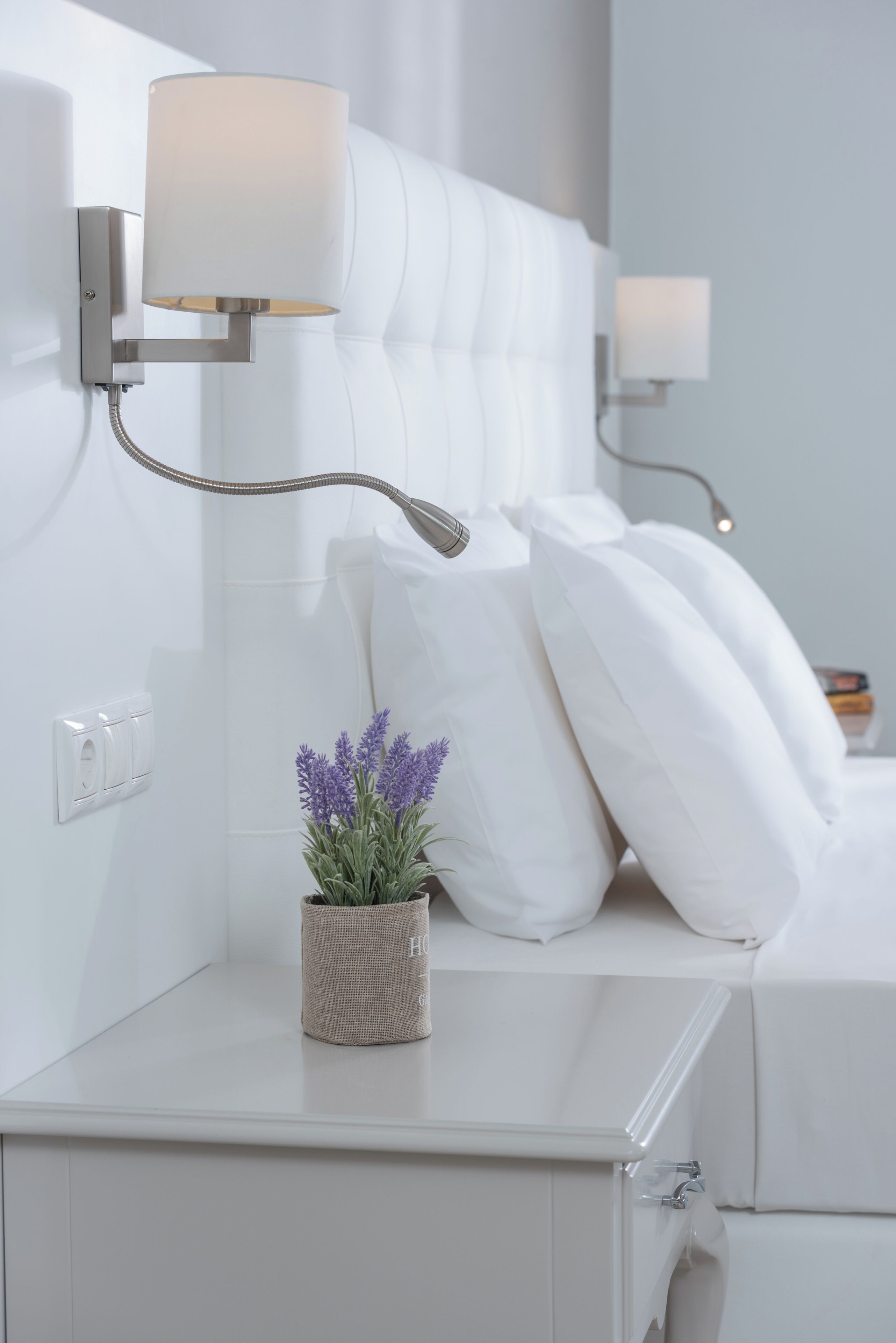 Un dormitorio moderno con cabecera blanca, lámpara de pared, una pequeña planta en maceta y una mesita de noche.