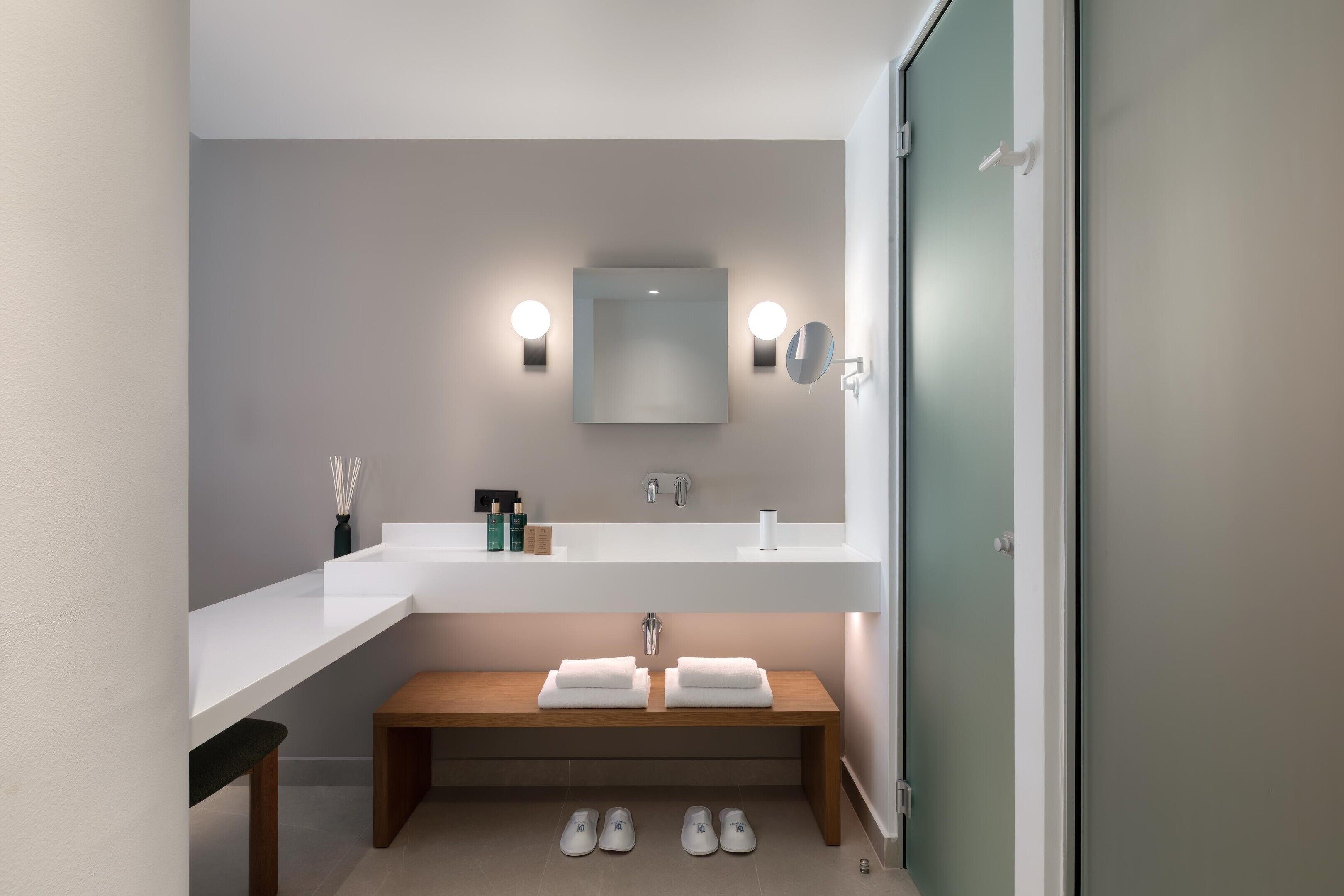 A modern bathroom with a white vanity, two sinks, a mirror, and a bench with towels.