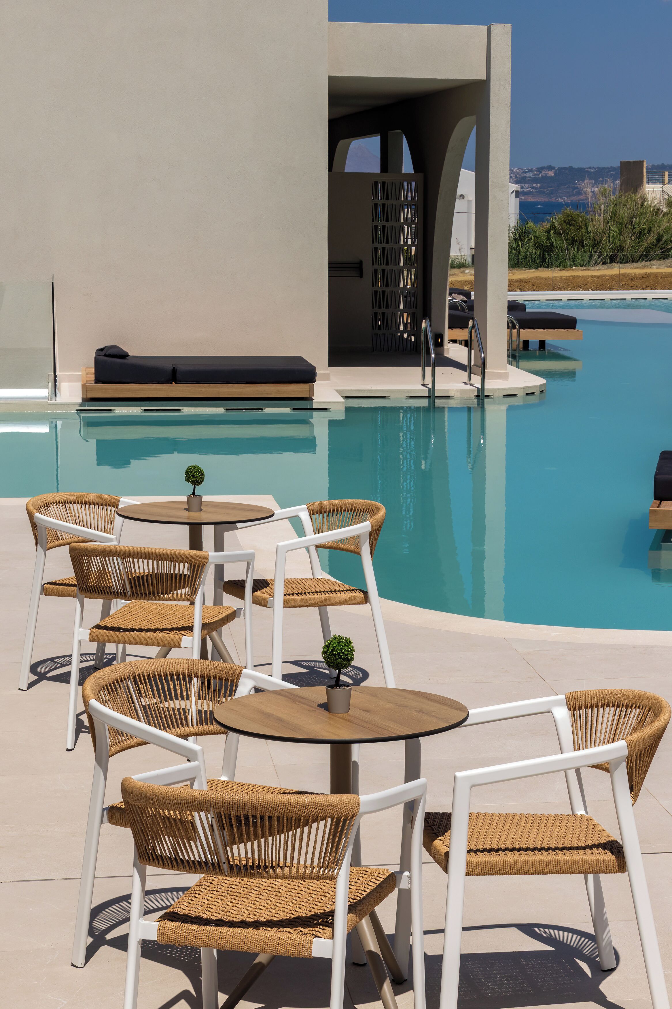 A poolside area with wicker chairs and tables, a small plant on one table, and a view of the sea in the background.