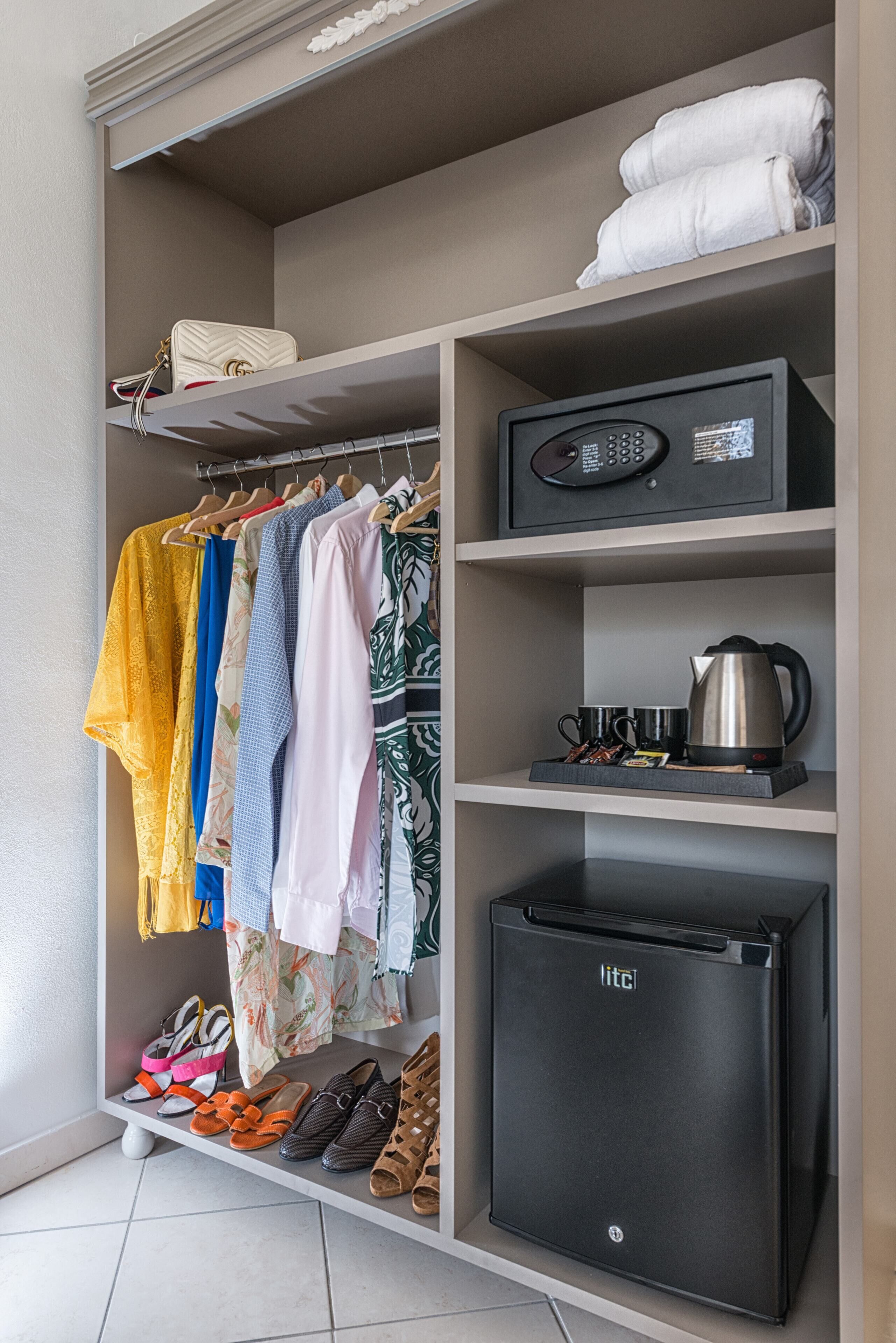 A hotel room with a wardrobe containing folded clothes, a microwave, a kettle, and a refrigerator.