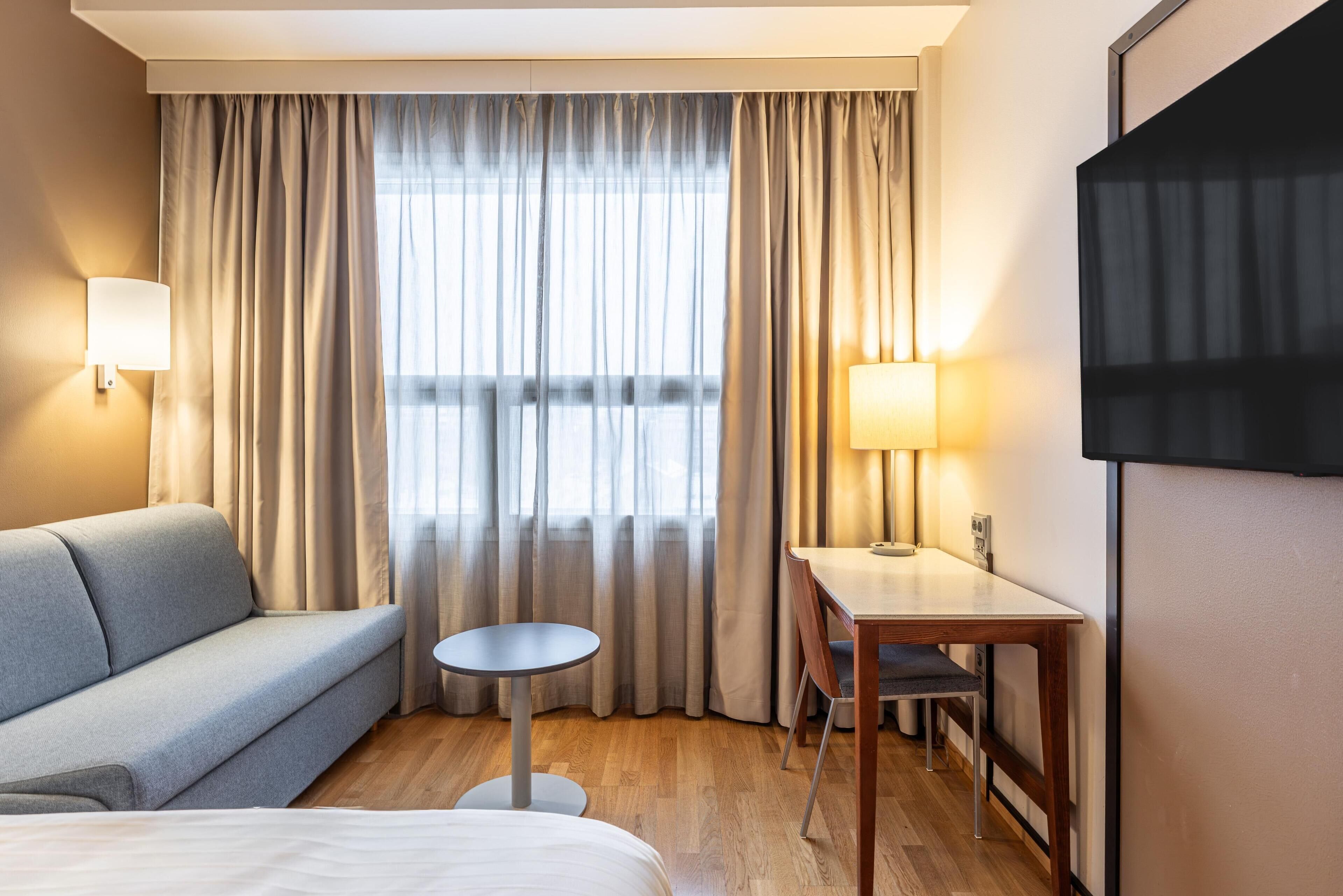 Une chambre d’hôtel équipée d’un canapé, d’une petite table ronde, d’un bureau avec une chaise et d’un téléviseur fixé au mur.