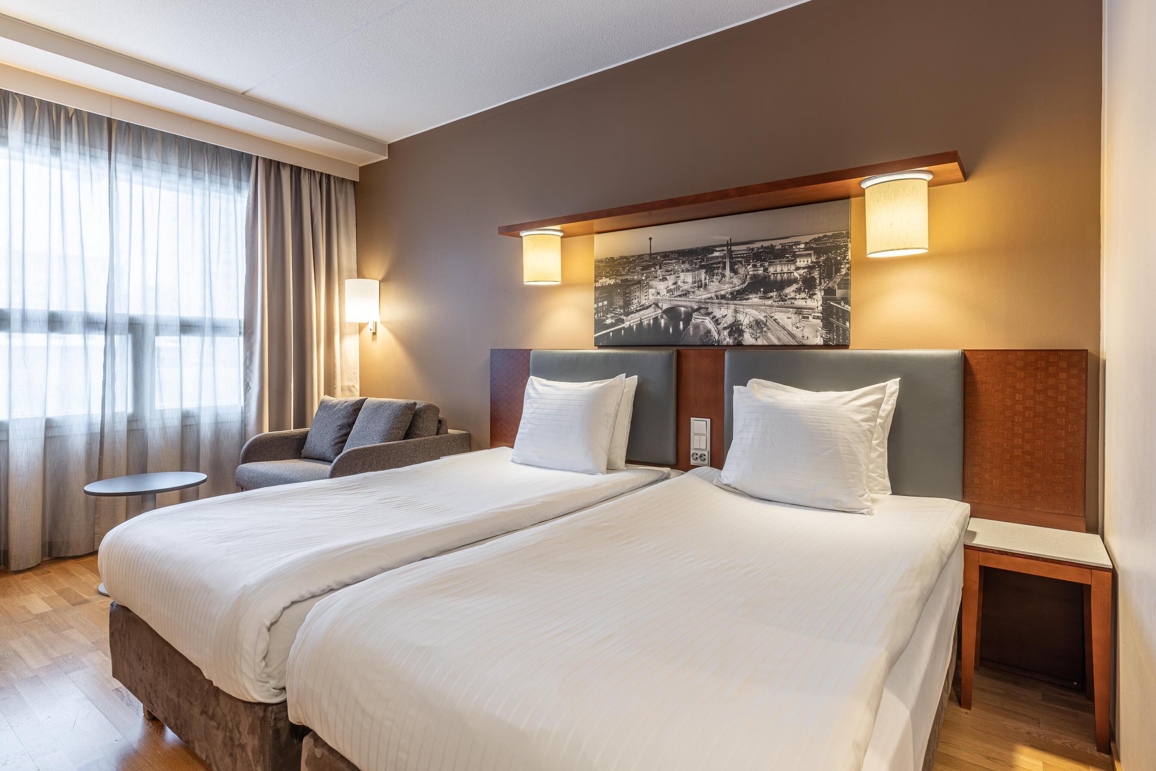 Une chambre d’hôtel avec deux lits, un canapé, une table ronde et un tableau représentant un paysage urbain accroché au mur.