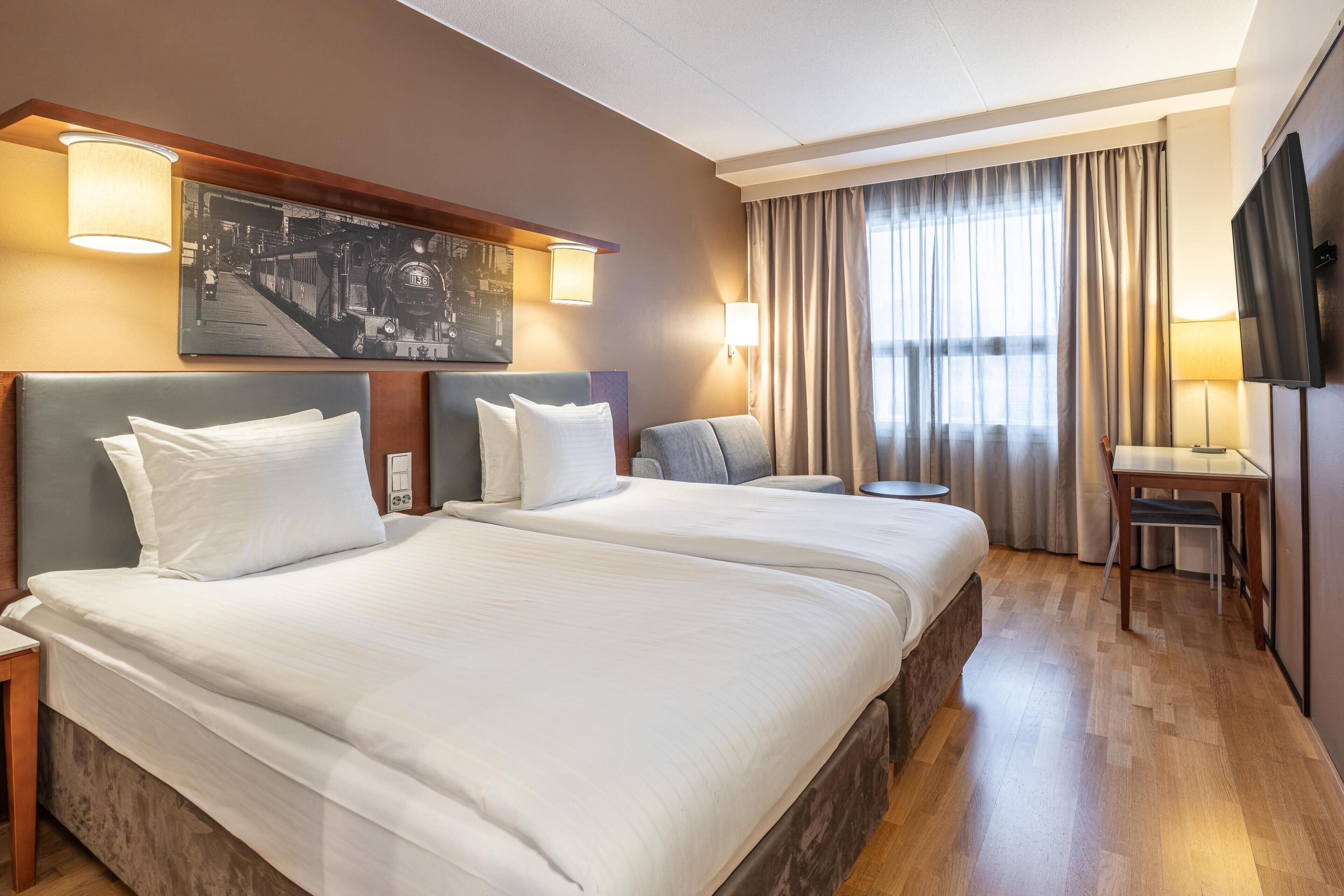 A hotel room with two beds, a desk, a chair, a television, and a large window with curtains.