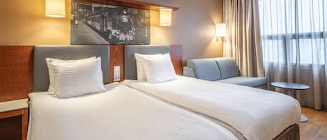 Quarto de hotel com duas camas, um sofá, uma mesinha e uma janela grande com cortinas.