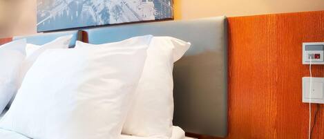 Hotellihuone, jossa on sänky, yöpöytä, seinälle asennettu ohjauspaneeli ja sängyn yläpuolella oleva kehystetty kuva.