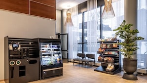 A modern hotel lobby with a self-service beverage station, seating area, and a large window offering a view of the city. - Scandic Postitalo (Tampere)