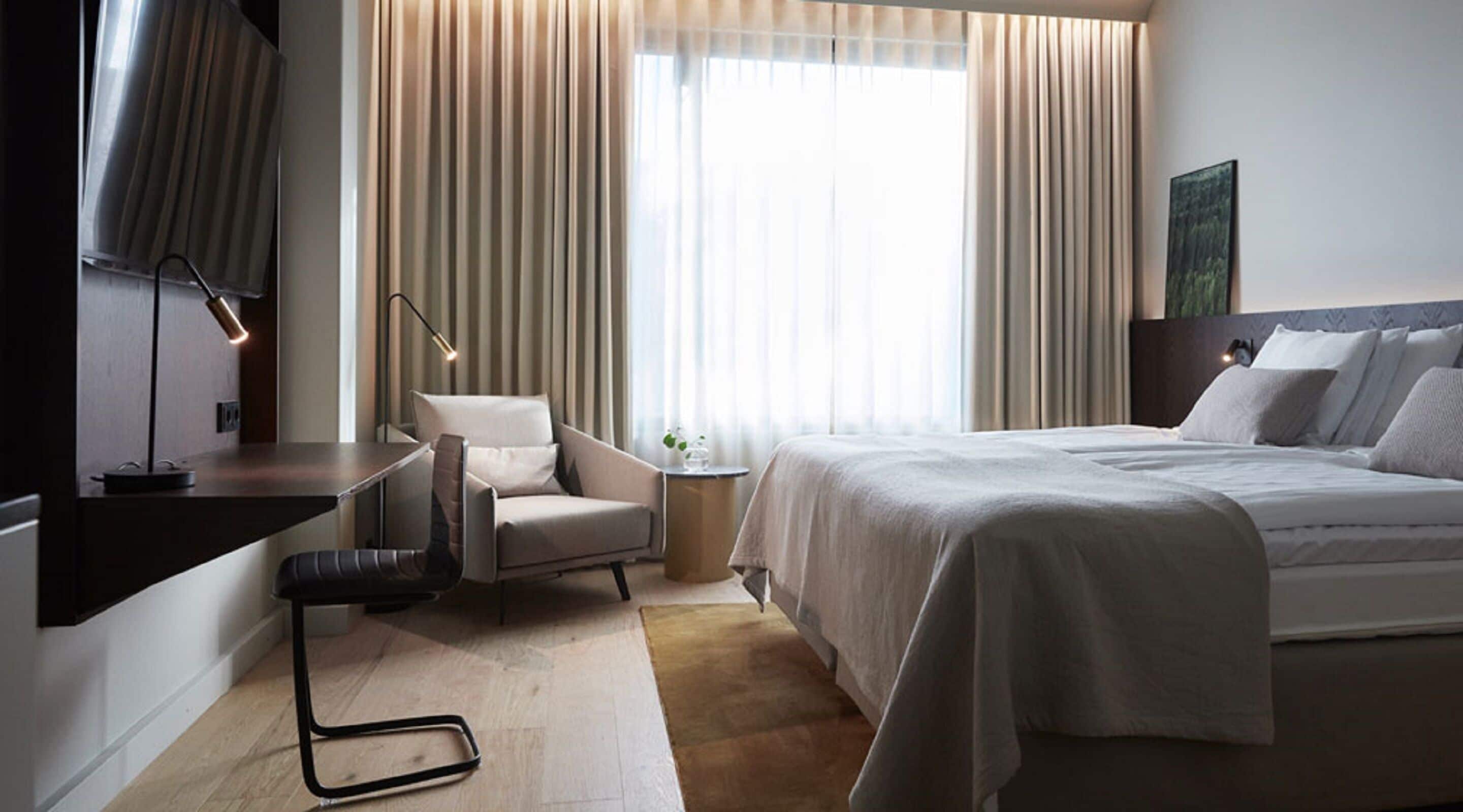 Une chambre d’hôtel moderne équipée d’un lit, d’une chaise, d’un bureau et d’une grande fenêtre avec des rideaux.