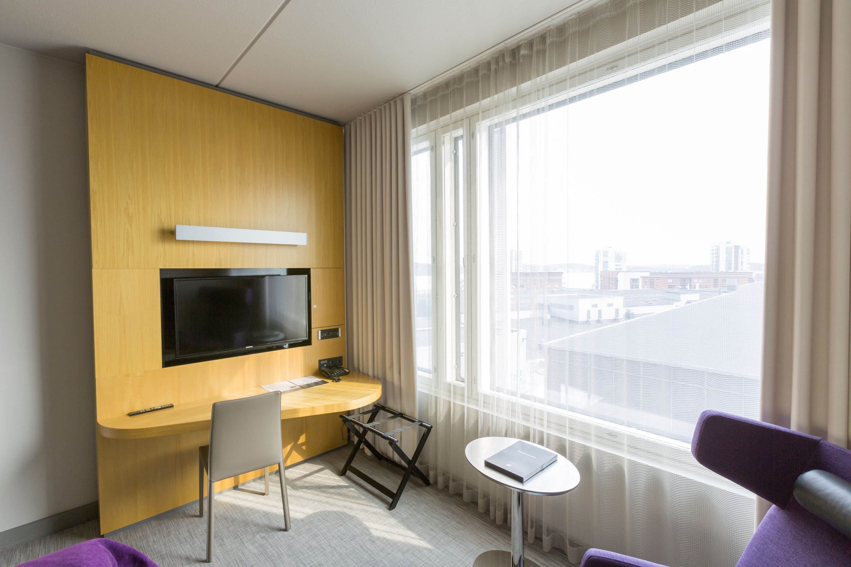 Habitación de hotel con escritorio de madera, televisor montado en la pared, una mesita y vistas de la ciudad a través de amplias ventanas.