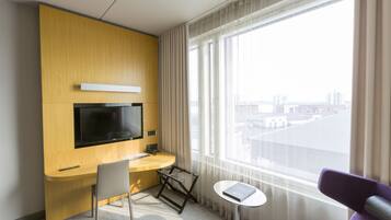 Habitación de hotel con escritorio de madera, televisor montado en la pared, una mesita y vistas de la ciudad a través de amplias ventanas.