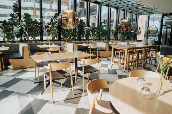 A modern indoor dining area with wooden tables and chairs, large windows, and indoor plants. - Solo Sokos Hotel Paviljonki (Jyväskylä)