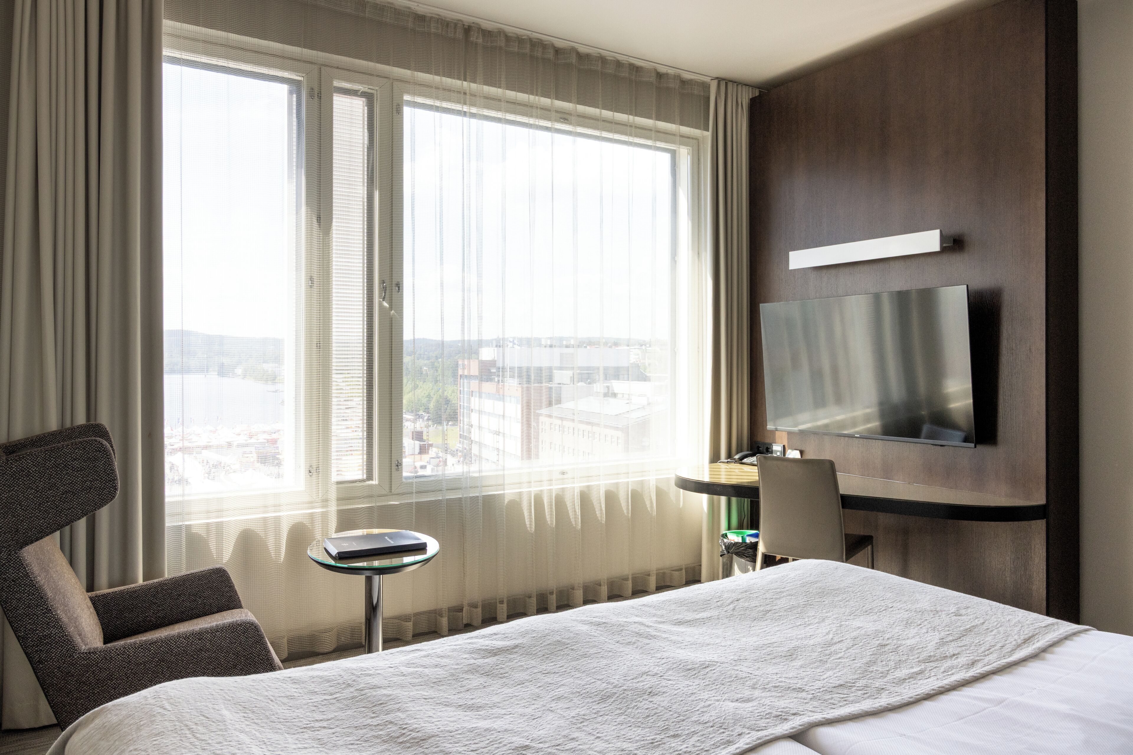 A hotel room with a bed, a desk with a chair, a television mounted on the wall, and a large window with curtains.