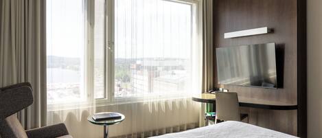 A hotel room with a bed, a desk with a chair, a television mounted on the wall, and a large window with curtains.