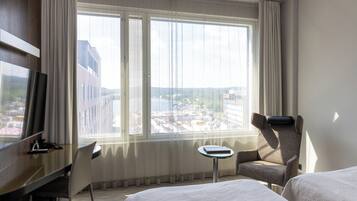 A hotel room with a large window, a desk, a chair, and two beds.
