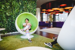 Une personne est assise sur un fauteuil vert agrémenté d’un coussin vert, entourée d’un mur verdoyant et d’un intérieur moderne avec des sièges violets et des suspensions lumineuses orange.