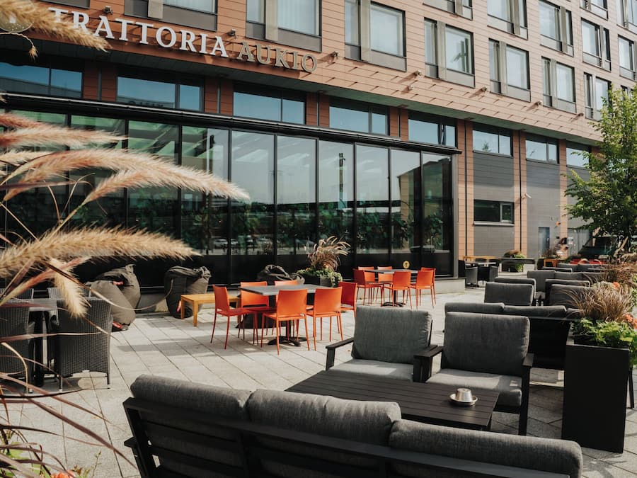 Et udendørs opholdsområde med borde og stole, en bygning med skiltet 'TRATTORIA AKIO' og store vinduer.