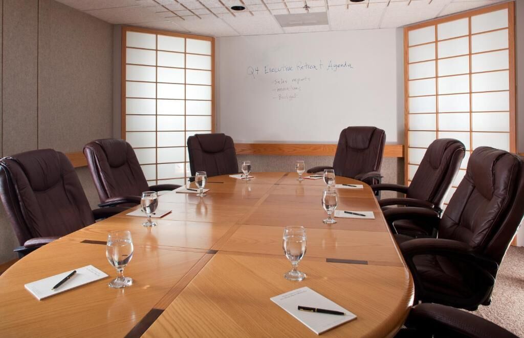 Uma sala de conferência com uma longa mesa de madeira, seis cadeiras de couro marrom e um quadro branco com tópicos da pauta.