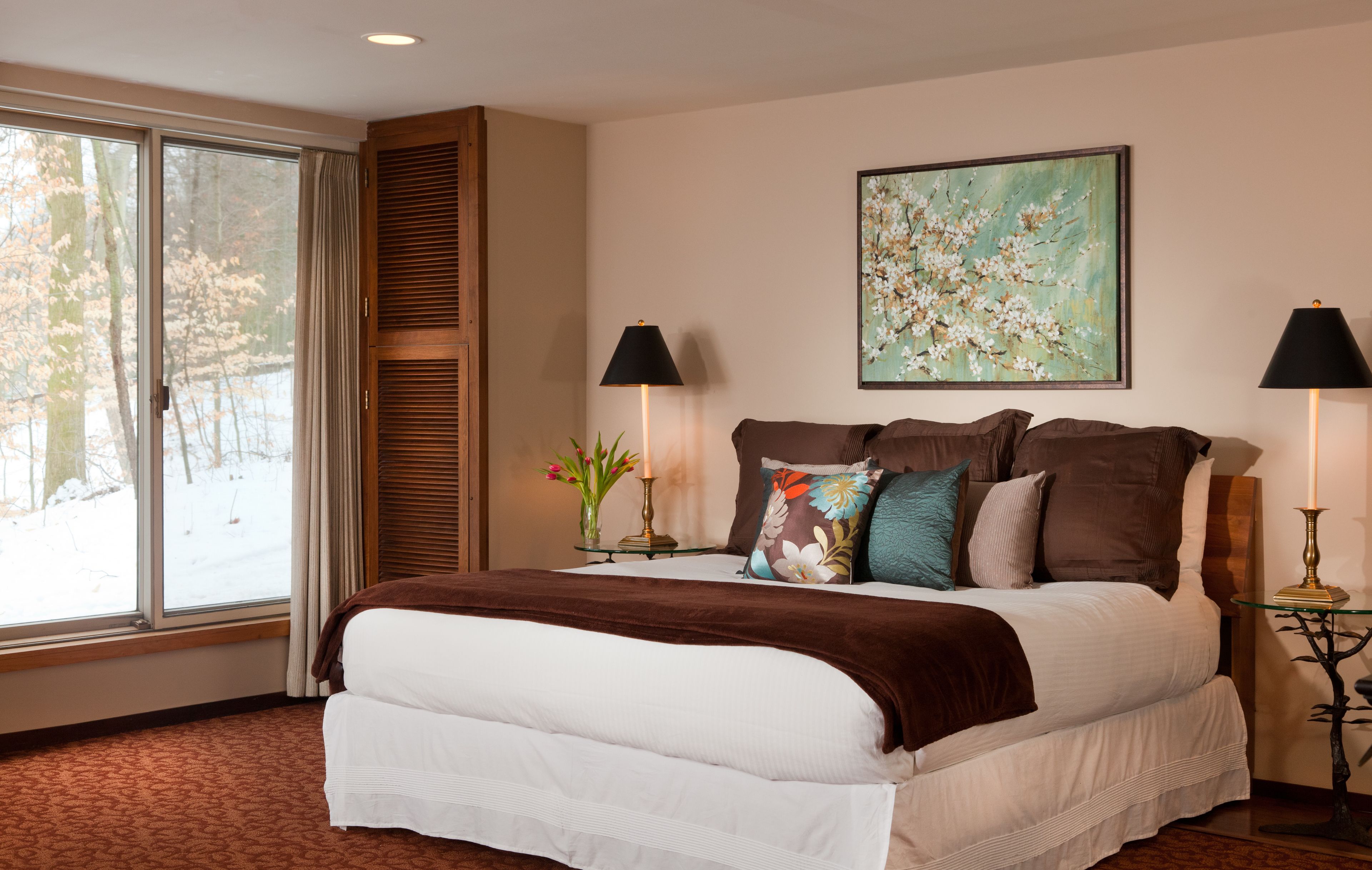 A bedroom with a large bed, two bedside lamps, a window with a view of trees, and a painting on the wall.