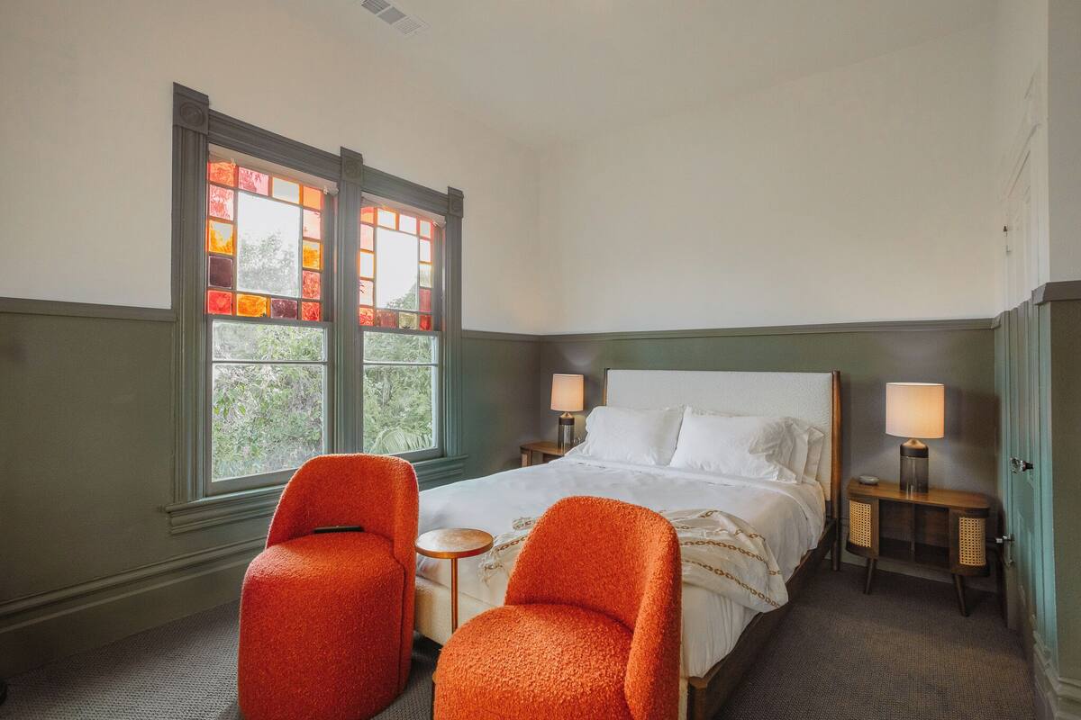 A hotel room with a large bed, two bedside tables with lamps, a small round table, two orange chairs, and a window with stained glass.
