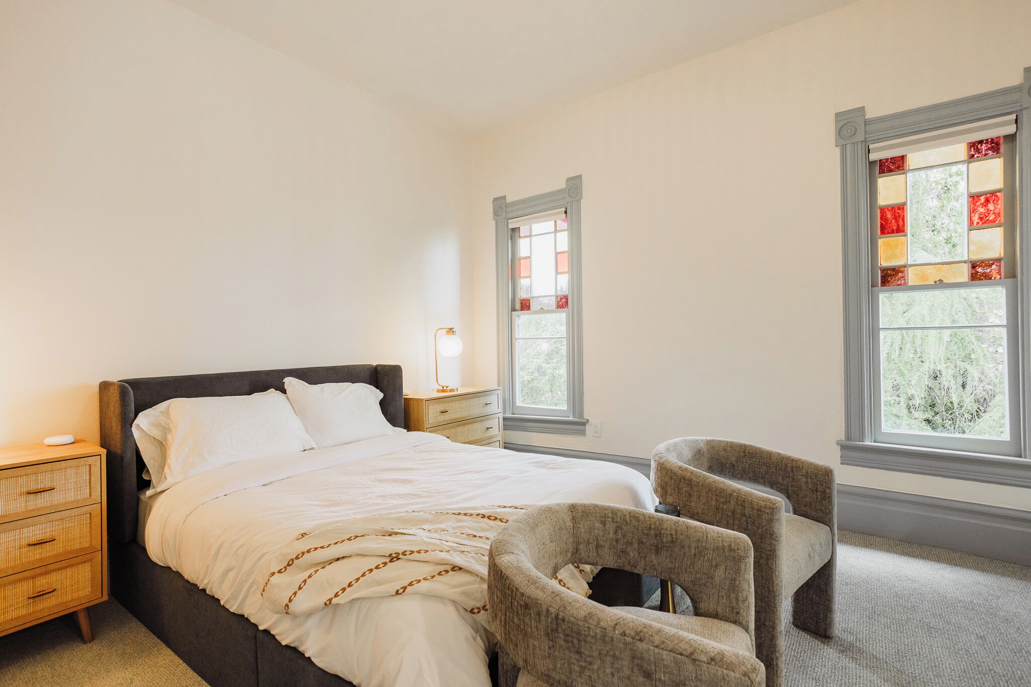 Un dormitorio con cama, dos sillas, una mesita de noche, una lámpara y dos ventanas con vidrieras.