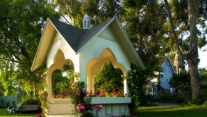 A gazebo with a white exterior and a peaked roof, surrounded by lush greenery and flowering plants. - Kaleidoscope Inn & Gardens (Nipomo)