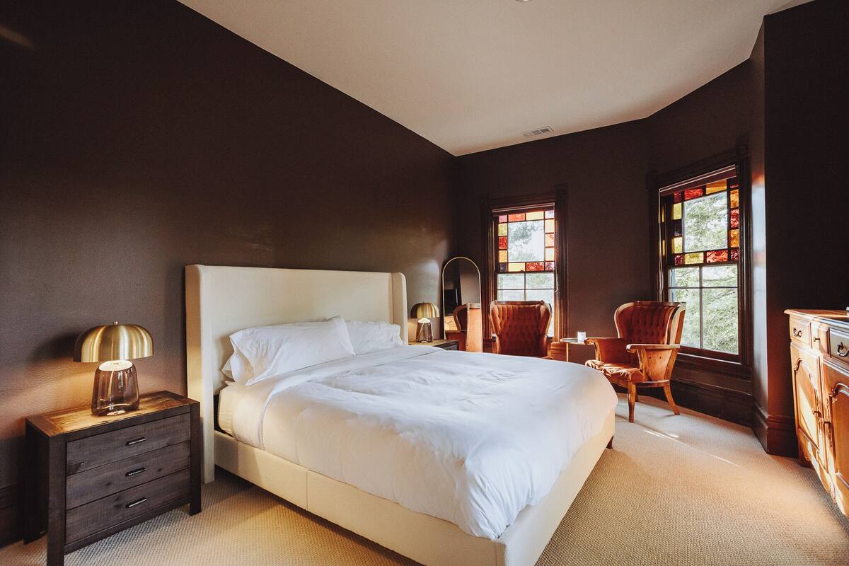 A bedroom with a large bed, a nightstand with drawers, a chair, a dresser, and two windows with stained glass.