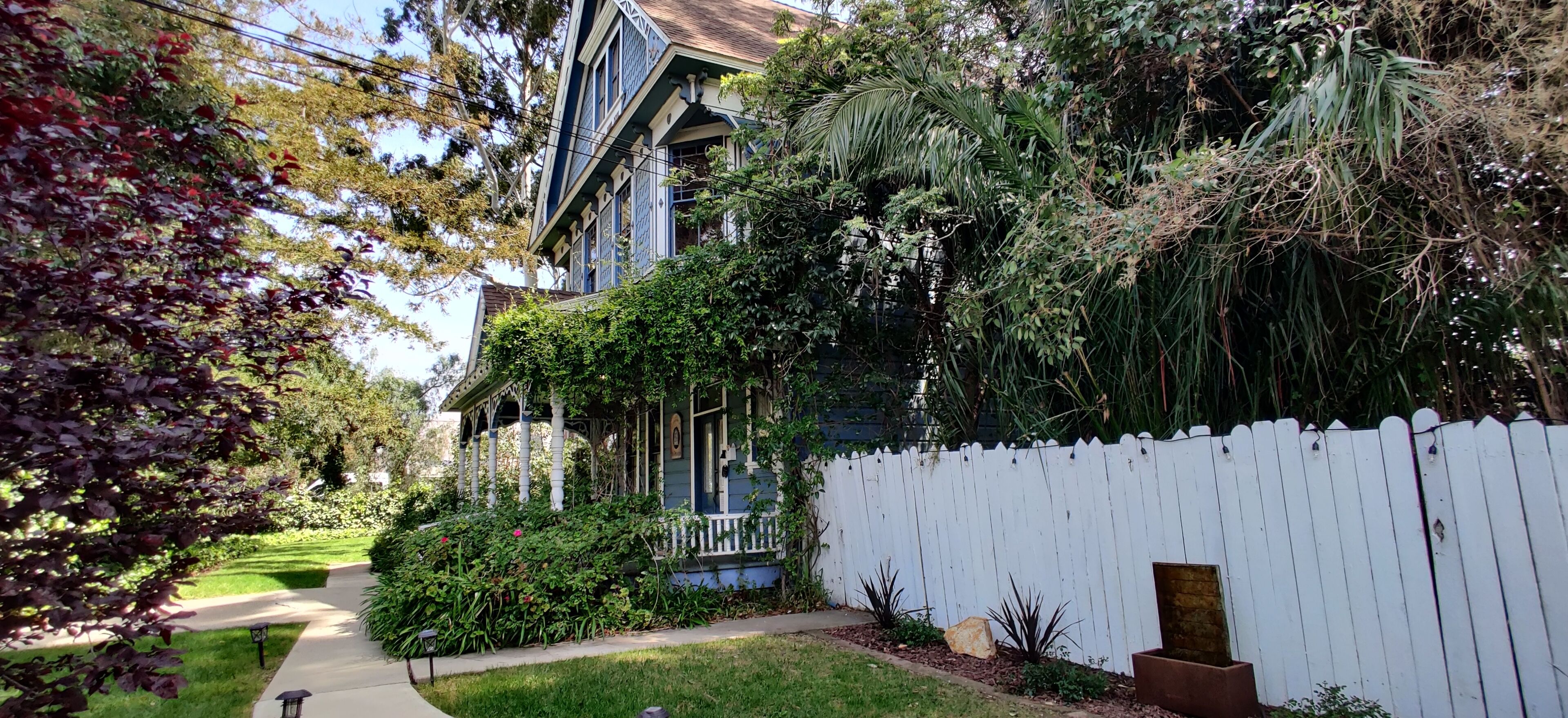 A two-story house with a porch, surrounded by a white picket fence, and lush greenery.