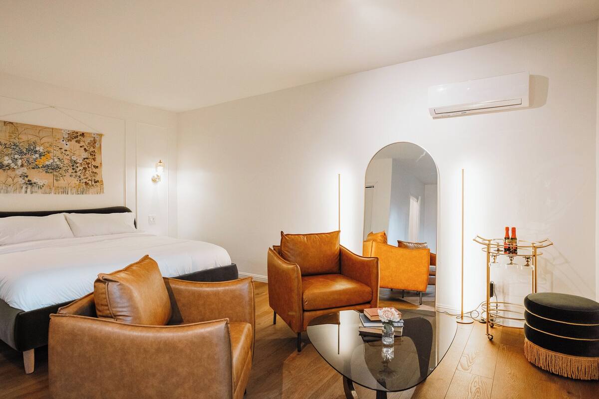 A modern hotel room with a large bed, leather armchairs, a glass coffee table, and a small side table with bottles.