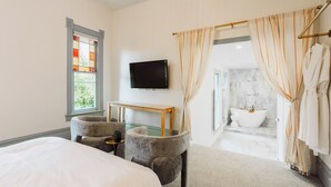 A bedroom with a bed, a desk, two armchairs, a TV, and a bathroom with a bathtub and shower. - Kaleidoscope Inn & Gardens (Nipomo)