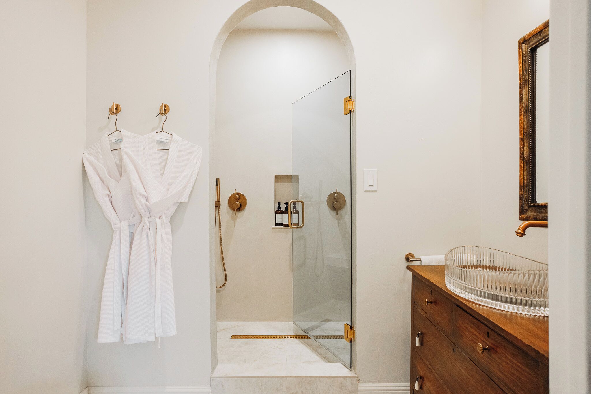 Une salle de bain avec une douche à parois de verre, un meuble-lavabo en bois avec un vasque en verre, et une robe accrochée au mur.