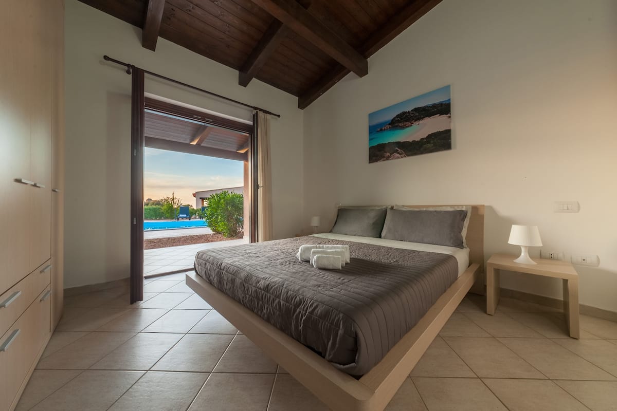 Ein Schlafzimmer mit Bett, Nachttisch, einem Gemälde und Blick auf Pool und Grünflächen.