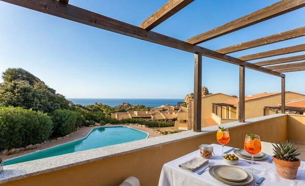 Ein Balkon mit Blick auf einen Swimmingpool und das Meer, ein Esstisch gedeckt und ein Blumentopf.