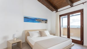 Une chambre à coucher comprenant un lit, une table de chevet, une lampe, un tableau représentant un paysage côtier et une porte-fenêtre coulissante donnant sur un balcon.
