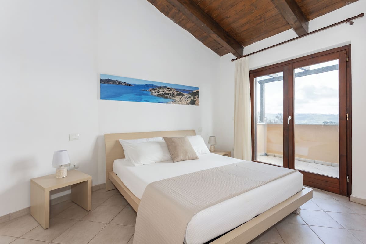 Ein Schlafzimmer mit einem Bett, einem Nachttisch, einer Lampe, einem Bild mit einer Küstenansicht und einer Glastür zum Balkon.
