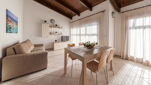 Un vaste espace de vie doté d’un plafond en bois, d’un canapé beige, d’une table à manger avec des chaises, d’un téléviseur posé sur un meuble blanc et d’un tableau représentant un paysage côtier.