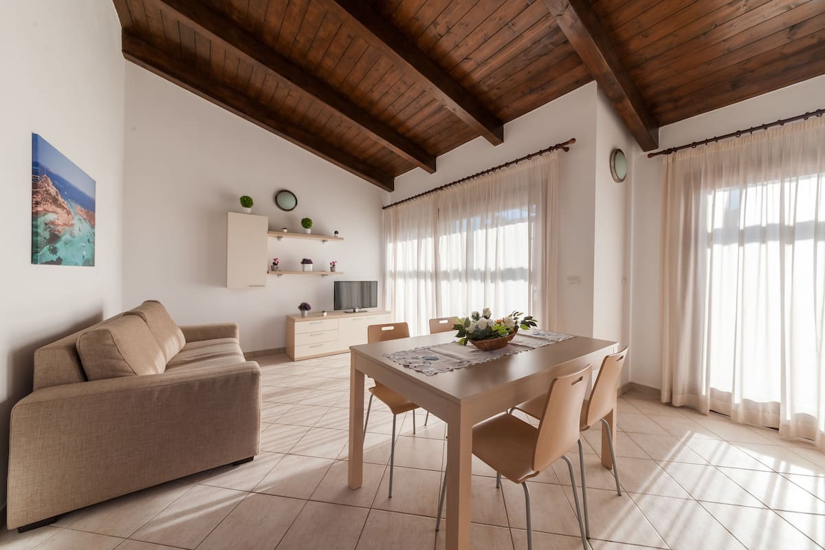 Ein geräumiger Wohnbereich mit Holzbalkendecke, einem beigefarbenen Sofa, einem Esstisch mit Stühlen, einem Fernseher auf einem weißen Schrank und einem Gemälde einer Küstenlandschaft.