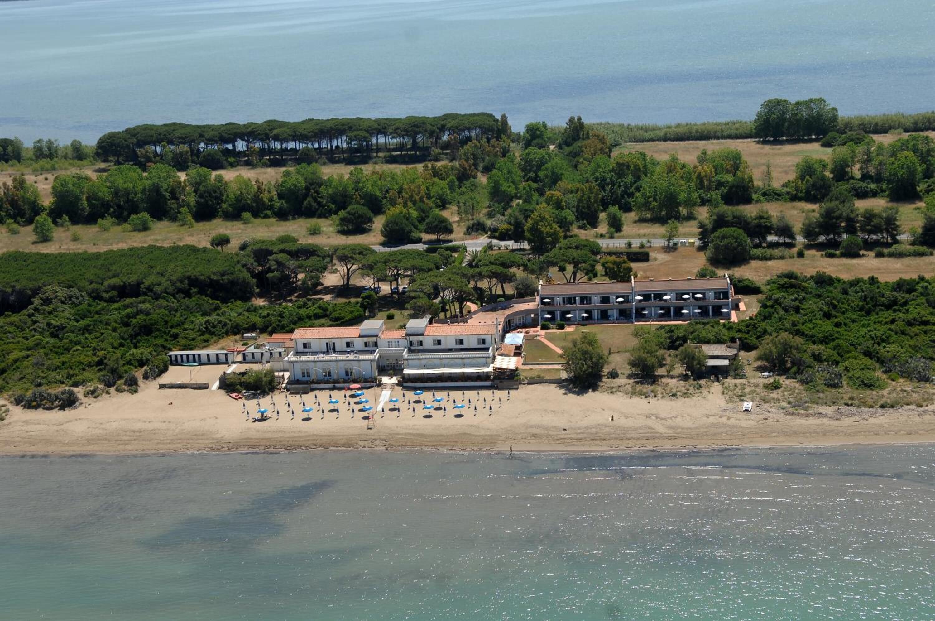 An aerial view of a coastal hotel with multiple buildings, a beach with umbrellas, and a forested area in the background.