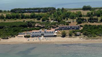 Vista aérea de un hotel costero con varias construcciones, una playa con sombrillas y un área boscosa al fondo.