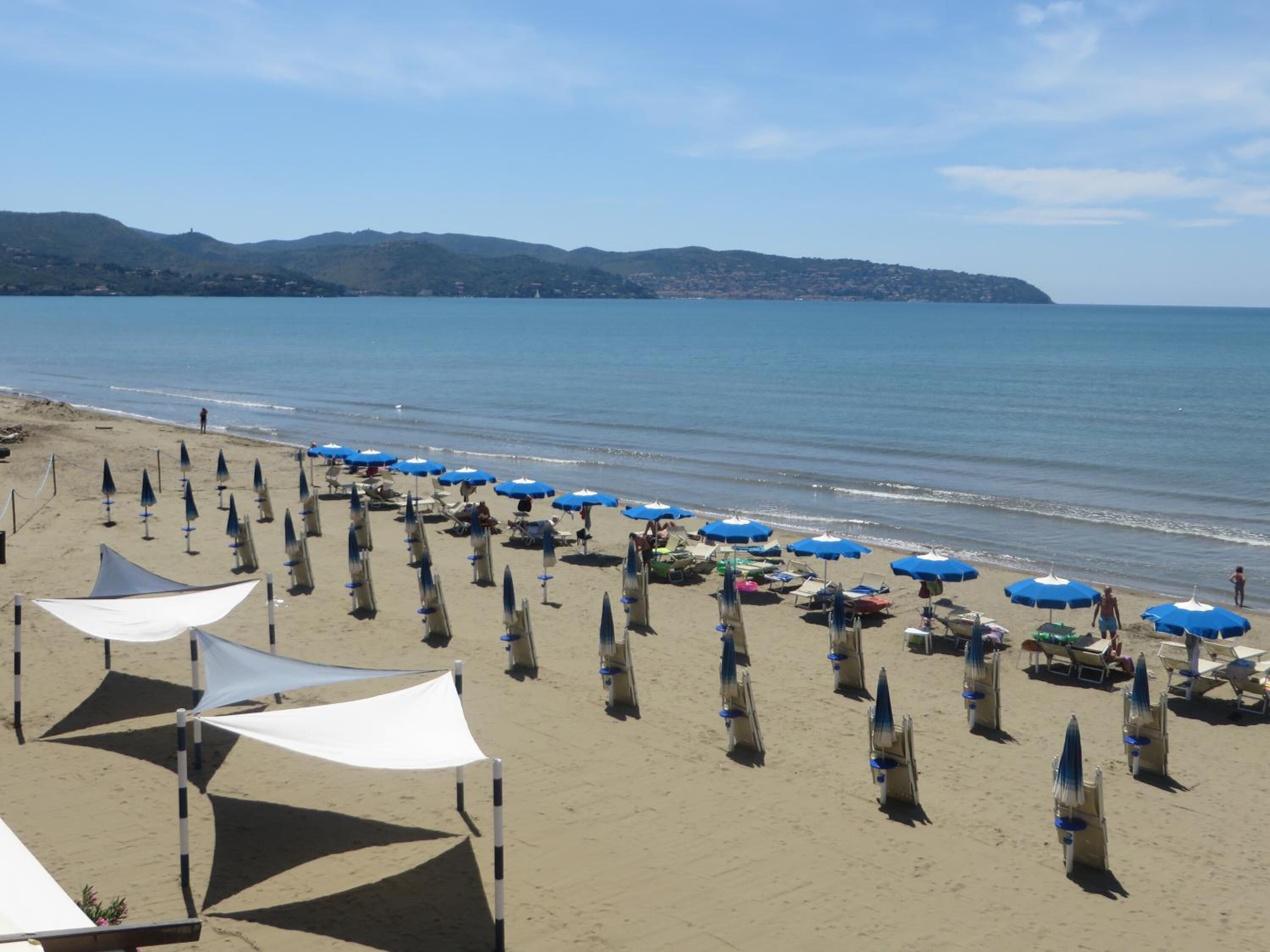 Una playa con hileras de sombrillas y sillas, cielo despejado y una vista distante de colinas.